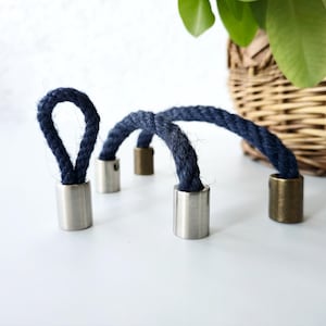 May include: A navy blue rope handle with metal and brass detailing. The rope is looped and attached to cylindrical metal pieces. The design is simple and modern, suitable for various home decor projects.