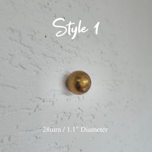 Może przedstawiać: Pojedynczy, okrągły, mosiężny uchwyt o średnicy 2,8 cm, zamontowany na teksturowanej białej powierzchni. Tekst "Style 1" jest napisany nad uchwytem. Tekst "28mm / 1.1" Diameter" jest napisany poniżej.