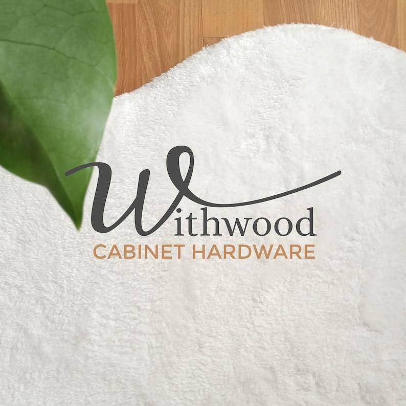 Może przedstawiać: Zbliżenie białego, puszystego dywanu z tekstem "Withwood CABINET HARDWARE" w ciemnoszarym i brązowym kolorze. Zielony liść jest widoczny w lewym g&oacute;rnym rogu, a tło to drewniana podłoga.