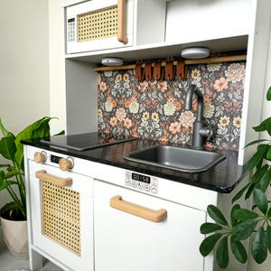 Può includere: Una cucina giocattolo in legno bianco con piano di lavoro nero, lavello in acciaio inossidabile e forno a microonde. La cucina presenta maniglie in legno chiaro e dettagli in canna sulla porta del forno. Il paraschizzi ha un motivo floreale.