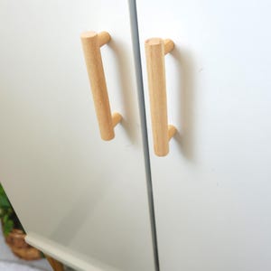 May include: Two wooden cabinet door pulls with a light brown finish. The pulls are mounted on a white cabinet door.
