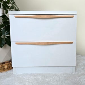Puede incluir: Mesita de noche blanca con dos cajones y tiradores de madera. La mesita de noche tiene un diseño simple y moderno.