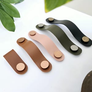 Könnte beinhalten: Fünf Leder-Schubladengriffe in verschiedenen Farben, darunter Braun, Rosa, Grün und Schwarz. Jeder Griff hat an jedem Ende einen runden Metallknopf.