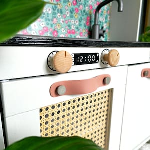 May include: White play kitchen with a black countertop, a digital clock that reads 12:00, and two wooden knobs. The oven door has a woven rattan panel and a pink leather handle with a silver rivet.