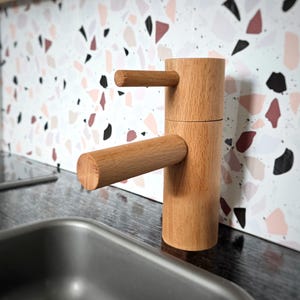 Puede incluir: Un grifo de cocina de madera con un cuerpo cilíndrico y dos manijas horizontales. El grifo está hecho de madera de color claro y está colocado contra un salpicadero con un patrón de terrazo en tonos de rosa, negro y blanco. Un fregadero de acero inoxidable está en primer plano.