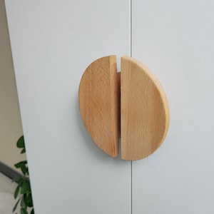 May include: Two wooden half-circle cabinet pulls, each with a natural wood finish, are attached to a white cabinet door.