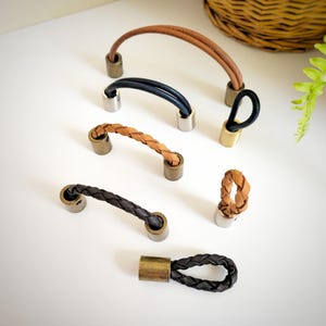 May include: A selection of leather drawer pulls and handles in different styles and colours. The collection includes braided and looped designs in brown, black, and tan, with metal accents. These hardware pieces are suitable for furniture and home decor projects.
