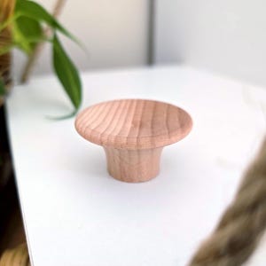 Könnte beinhalten: Ein hellfarbiger Holzschrankknopf mit abgerundeter Oberseite und ausgestellter Basis. Der Knopf besteht aus Naturholz und hat sichtbare Holzmaserung. Der Knopf liegt auf einer weißen Oberfläche.