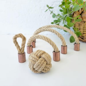 May include: A collection of nautical-themed decorative items. The set includes a rope knot, a rope loop, and rope handles with copper-colored metal ends. The items are arranged on a white surface, with a wicker basket and greenery in the background.