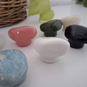 Può includere: Una collezione di pomelli per mobili in ceramica di vari colori, tra cui rosa, bianco, nero, verde e blu. I pomelli hanno una forma arrotondata, a fungo, e sono disposti su una superficie bianca. Sullo sfondo è visibile un cestino intrecciato.