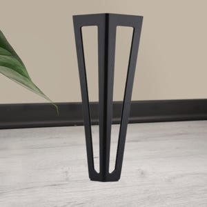 May include: A single, black metal furniture leg with a triangular design. The leg has a tapered shape with three rectangular cutouts. It stands on a light gray wooden floor, with a neutral-colored wall in the background.