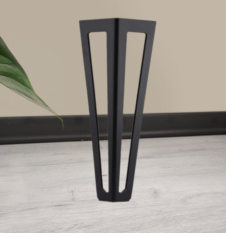 May include: A single, black metal furniture leg with a triangular design. The leg has a tapered shape with three rectangular cutouts. It stands on a light gray wooden floor, with a neutral-colored wall in the background.