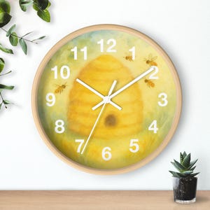 May include: A round wall clock with a light wood frame featuring a watercolor-style illustration of a yellow beehive and several small bees on a soft yellow background. The clock face displays large white numbers and white hour and minute hands.