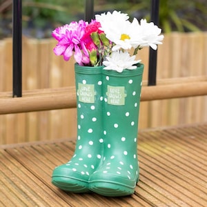 May include: Green wellington boots with white polka dots, holding pink and white flowers. The boots display the text "LOVE GROWS HERE" and are set on a wooden deck, creating a charming garden display.