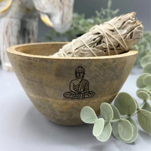 May include: A wooden bowl with a Buddha image engraved on the bottom. The bowl is filled with white sage smudge sticks.