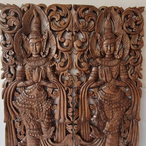 May include: A pair of rectangular dark brown wooden wall panels featuring intricate hand-carved relief designs of two identical figures in traditional attire with pointed headdresses, standing with hands pressed together in a prayer gesture.