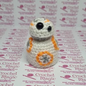 May include: A handmade crochet BB-8 droid figure. The figure is primarily white with gray and orange accents. The eyes are black. The base has a gray and orange star design. The background features the text "Crochet Magic".