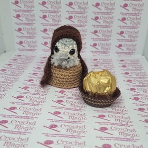 May include: A handmade crochet owl figurine with a brown hood and a tan body. The owl has a fluffy gray face and black eyes. Next to the owl is a wrapped chocolate candy in a brown paper cup.