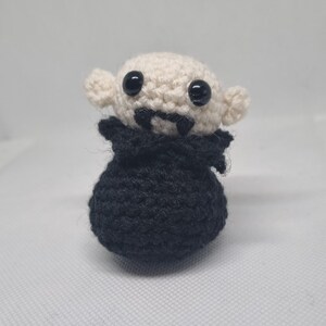 May include: A handmade crochet vampire doll with a cream-colored head, black eyes, and a black cape. The doll has small, pointed ears and two small fangs. The body is black and rounded, giving it a cute, cartoonish appearance.