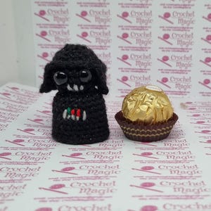 May include: A handmade black crochet figure with large eyes and a small mouth, next to a gold-wrapped chocolate candy. The crochet figure has a small design with red, white, and green accents. The background features a repeating pattern with the words "Crochet Magic".