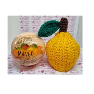 May include: A bath bomb and a crocheted mango. The bath bomb is round with a label that reads "Mango" and "Relax & Revive". The crocheted mango is yellow with a green leaf and brown stem. The background has the words "Crochet Magic".
