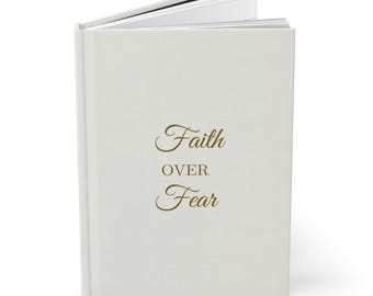May include: A cream-colored journal with the words "Faith Over Fear" in elegant gold script. The journal is open, revealing the white pages inside. The cover has a smooth texture.