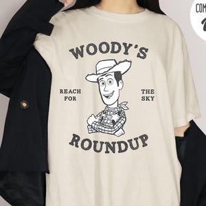 Pu&ograve; includere: T-shirt color avorio con grafica del cartone animato Woody e la scritta "WOODY'S ROUNDUP REACH FOR THE SKY". La maglietta &egrave; di Comfort Colors.