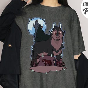 May include: Dark gray t-shirt featuring a wolf pack graphic. The design includes a large wolf with red eyes, a howling wolf in front of a blue moon, and several smaller wolves. The shirt has the text "COMFORT COLORS Pepper".