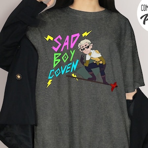 Puede incluir: Camiseta Comfort Colors gris oscuro con el texto "SAD BOY COVEN" en rosa, verde y azul, con rayos amarillos. La camiseta presenta una ilustraci&oacute;n de dibujos animados de un personaje con gafas de sol.