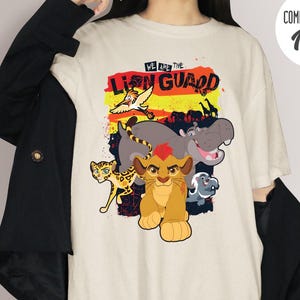 Puede incluir: Camiseta marfil Comfort Colors con un gráfico colorido de personajes de El Guardián del León. El diseño incluye un cachorro de león, un hipopótamo, un guepardo y otros animales, con el texto "WE ARE THE LION GUARD".