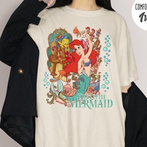May include: Ivory-colored t-shirt featuring a colorful graphic of Ariel from The Little Mermaid. The design includes Ariel with red hair and a teal tail, along with Flounder and floral elements. The shirt is labeled "Comfort Colors" and "Ivory".