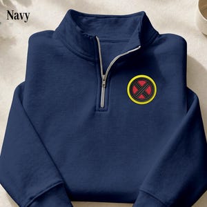 May include: A navy blue quarter-zip sweatshirt featuring a stand-up collar and a silver-toned zip. On the left chest, there is a circular embroidered patch with a yellow border, a black background, and a red X-shaped design in the centre.