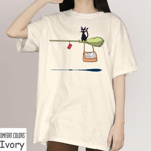 May include: Ivory Comfort Colors t-shirt featuring a whimsical illustration. A black cat sits on a broom with a sign that reads "NOT IN SERVICE." A small red object hangs from the broom. The shirt is a solid ivory color.
