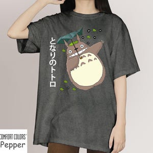 May include: Dark grey t-shirt with a cartoon Totoro character holding an umbrella. The Japanese text "となりのトトロ" is printed vertically on the left. The shirt is a Comfort Colors Pepper tee.