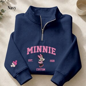 May include: A navy blue quarter-zip sweatshirt featuring a pink graphic of Minnie Mouse. The text reads MINNIE EST. 1928 CUSTOM. A small pink and white balloon graphic shaped like mouse ears is printed on the right sleeve near the cuff.