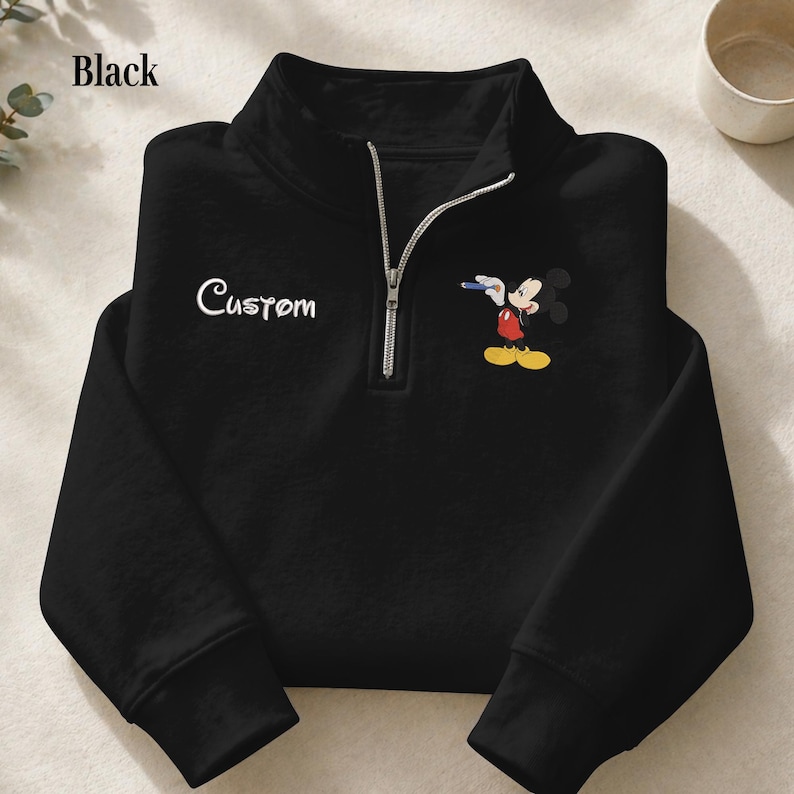 Puede incluir: Sudadera negra con cremallera de cuarto de cuello con texto "Custom" blanco y un gr&aacute;fico de Mickey Mouse. El personaje de dibujos animados es rojo, amarillo y negro. La sudadera tiene una cremallera plateada y un cuello alto.