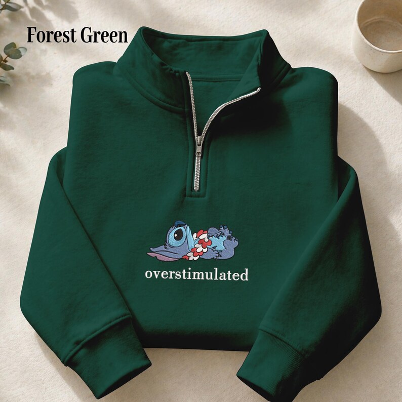 May include: A forest green quarter-zip sweatshirt featuring a silver zipper and a graphic of a blue character wearing a red and white flower lei. Below the character, the word overstimulated is printed in white, lowercase, serif font.