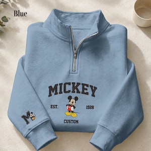 May include: A light blue quarter-zip sweatshirt featuring a graphic of Mickey Mouse. The text reads MICKEY EST. 1928 CUSTOM. A large letter M with a small Mickey Mouse graphic is printed on the left sleeve. The sweatshirt has a silver-toned zipper.