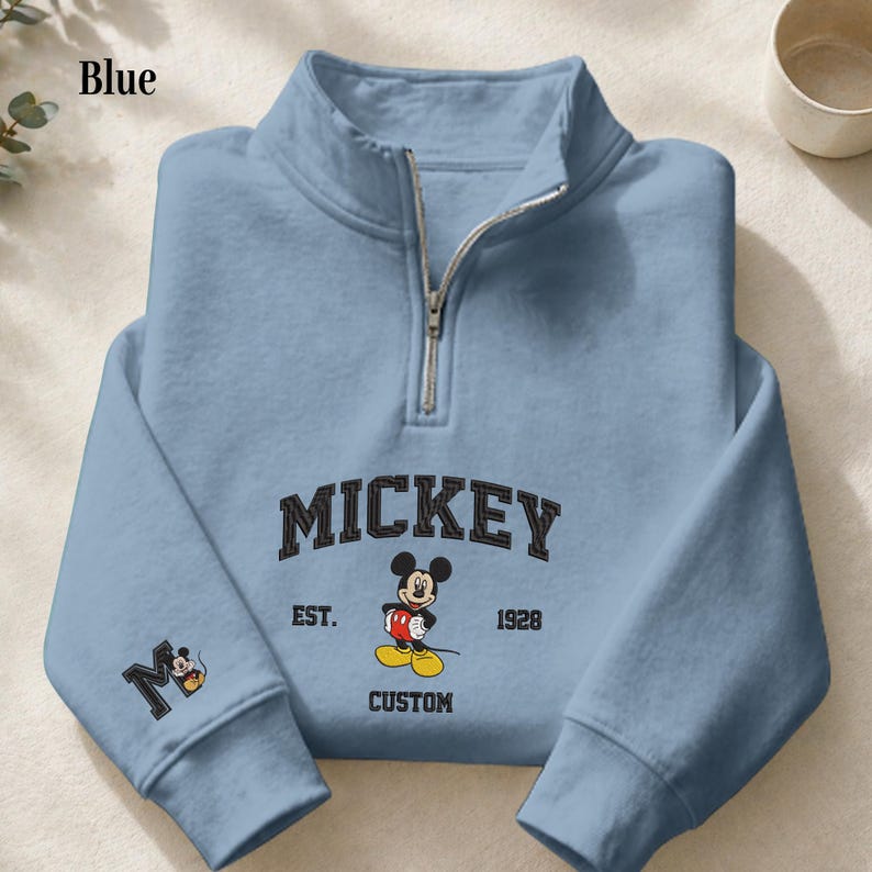 May include: A light blue quarter-zip sweatshirt featuring a graphic of Mickey Mouse. The text reads MICKEY EST. 1928 CUSTOM. A large letter M with a small Mickey Mouse graphic is printed on the left sleeve. The sweatshirt has a silver-toned zipper.