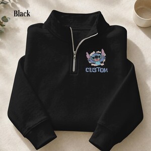 May include: Black quarter-zip sweatshirt featuring a cartoon character design. The design includes a blue cartoon character bursting through a hole, with the word "CUSTOM" below it. The sweatshirt has a silver zipper.