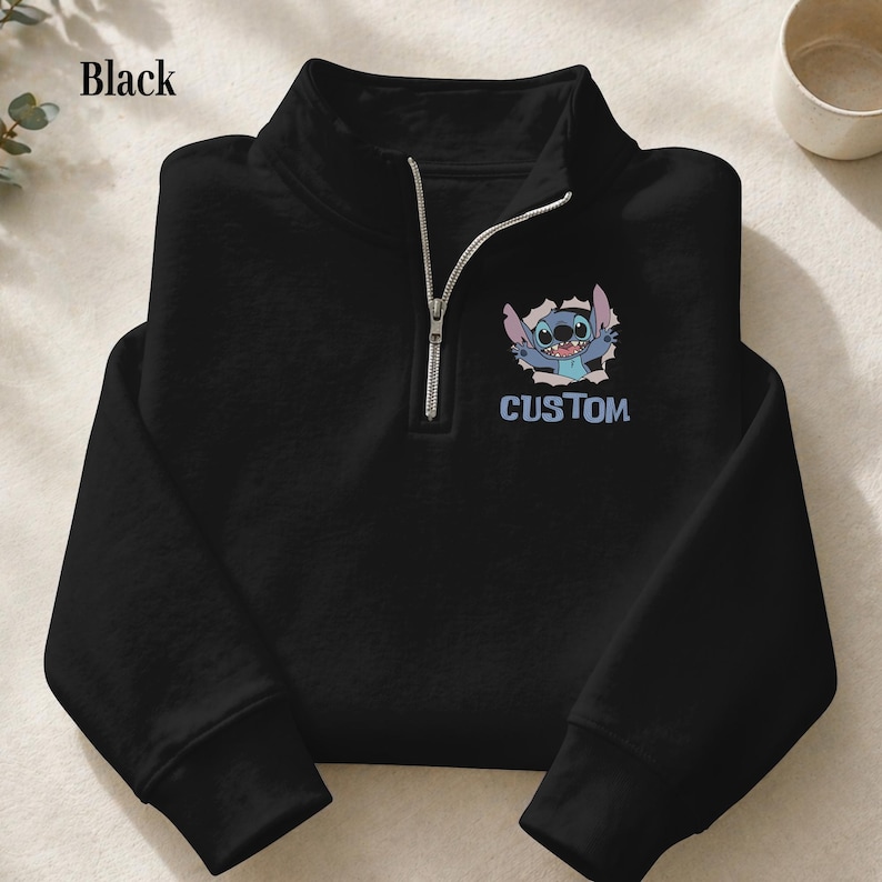 May include: Black quarter-zip sweatshirt featuring a cartoon character design. The design includes a blue cartoon character bursting through a hole, with the word "CUSTOM" below it. The sweatshirt has a silver zipper.