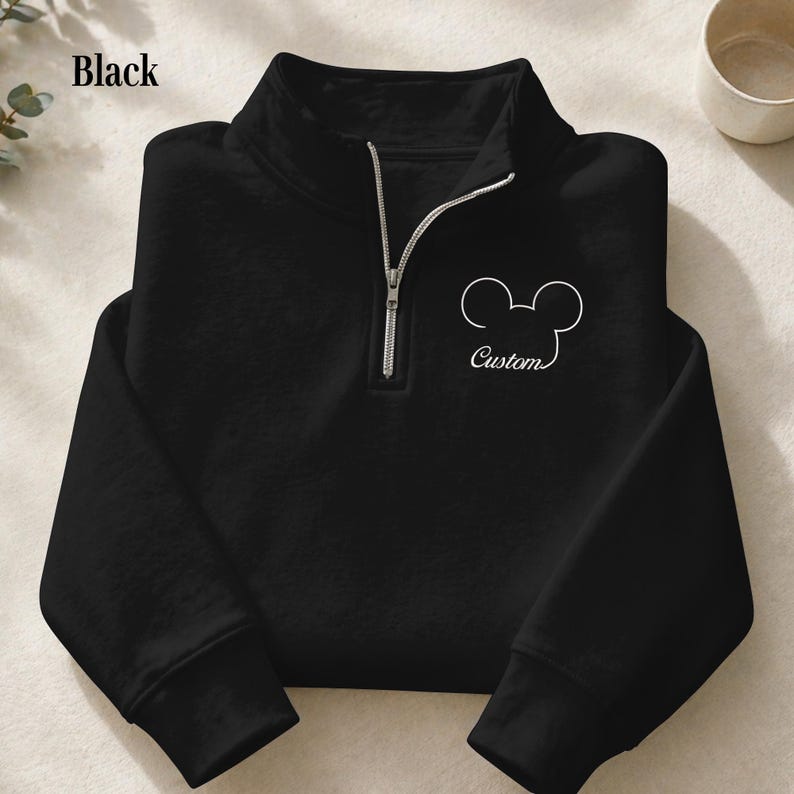 May include: Black pullover sweatshirt with a quarter-zip neckline. Features a white outline of a mouse head and the word "Custom" in script. The sweatshirt is a solid black color.