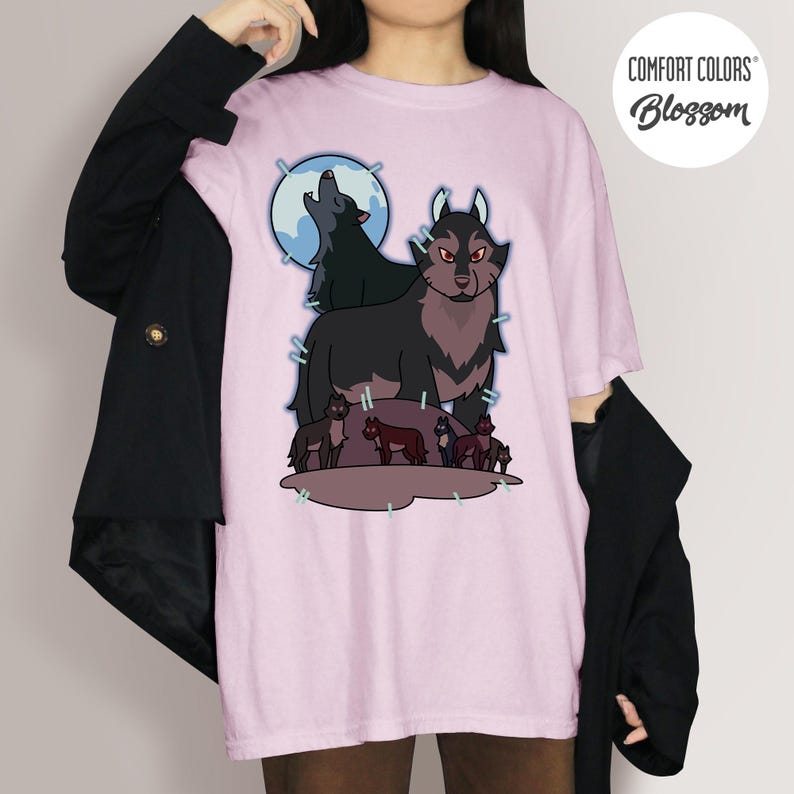 May include: A light pink t-shirt featuring a wolf pack illustration. The design includes a large wolf with red eyes, smaller wolves, and a howling wolf in front of a blue moon. The shirt has the text "COMFORT COLORS" and "Blossom" in a circle.