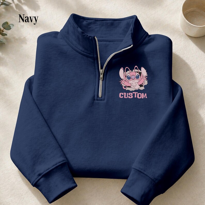 May include: Navy blue quarter-zip sweatshirt featuring a pink cartoon character with the word "CUSTOM" printed below. The sweatshirt has a silver zipper and a high collar. The word "Navy" is printed in the upper left corner.