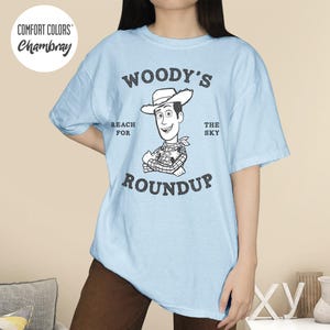 Pu&ograve; includere: T-shirt azzurra con una grafica di Woody di Toy Story. Il design include il testo "Woody's Roundup" con l'immagine di Woody e le frasi "Reach for" e "The Sky". La maglietta ha maniche corte e girocollo.