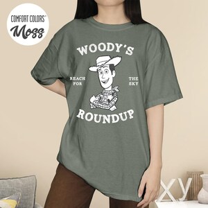 Pu&ograve; includere: Una maglietta verde muschio con una grafica di Woody di Toy Story. Il design include il testo "WOODY'S ROUNDUP" con "REACH FOR THE SKY" sopra il personaggio. La maglietta ha una vestibilit&agrave; ampia e maniche corte.
