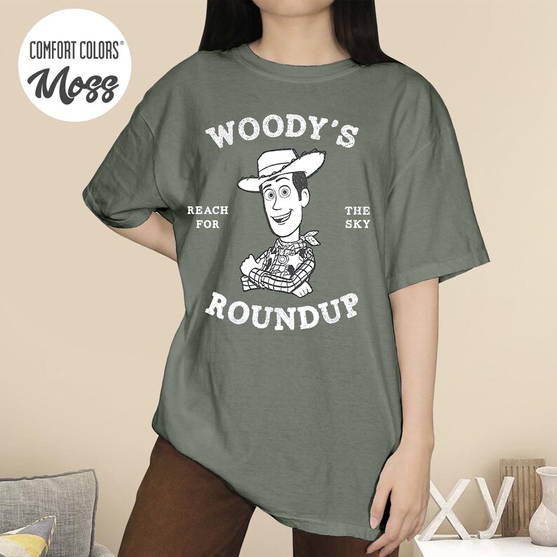 Pu&ograve; includere: Una maglietta verde muschio con una grafica di Woody di Toy Story. Il design include il testo "WOODY'S ROUNDUP" con "REACH FOR THE SKY" sopra il personaggio. La maglietta ha una vestibilit&agrave; ampia e maniche corte.