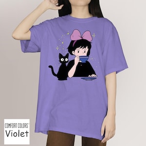 May include: Violet Comfort Colors t-shirt featuring a cartoon illustration of a girl with a pink bow, drinking from a blue cup, and a black cat. The graphic is set against a purple background. The shirt has a relaxed fit.