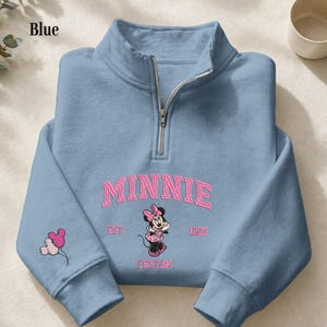 May include: A light blue quarter-zip sweatshirt featuring a pink embroidered Minnie Mouse graphic on the chest with the text MINNIE EST. 1928 CUSTOM. The left sleeve displays a small pink and light blue balloon graphic shaped like mouse ears.