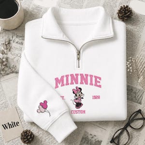May include: A white quarter-zip sweatshirt featuring pink embroidered text that reads MINNIE and CUSTOM, with the year 1928. The center displays a pink Minnie Mouse graphic, while the sleeve features a small design of three pink balloons.